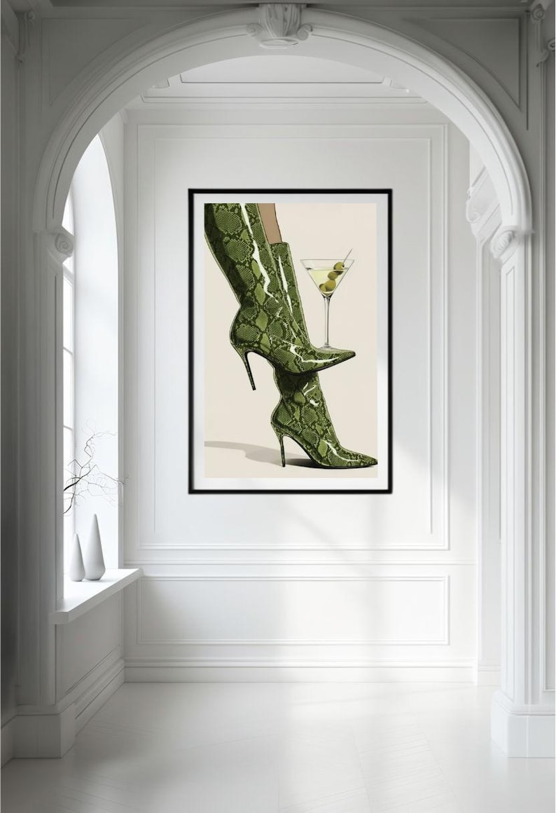 Op de afbeelding: Illustratie van groene stiletto laarzen met slangenprint en een martini glas. De laarzen zijn het middelpunt, met een hoge hak en spitse neus. Het martini glas bevat olijven. Het kunstwerk is zwart omlijst.