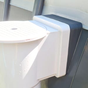 Könnte beinhalten: Eine weiße Regenwassertonne aus Kunststoff mit perforiertem Deckel und einer dunkelgrauen Halterung. Die Tonne ist zur Regenwassergewinnung an einer grauen Oberfläche, wahrscheinlich einer Außenwand, befestigt.