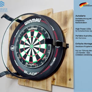 Può includere: Un bersaglio per freccette Winmau nero con un bersaglio verde e rosso, montato su assi di legno. È dotato di una luce LED circostante e del testo "BLADE 6". L'immagine include anche il testo "Autodarts" e "Made in Germany".