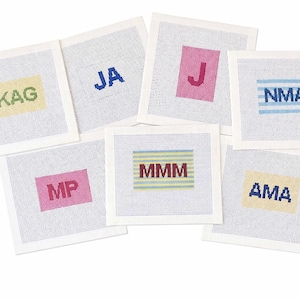 May include: A collection of square needlepoint coasters with various initials. Each coaster features a different initial or set of initials in a contrasting color, such as blue, pink, or green, set against a neutral background. The coasters are arranged on a white surface.