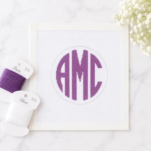 Puede incluir: Un lienzo cuadrado para bordar que presenta un monograma morado con las letras AMC centradas dentro de un contorno circular. Se incluyen dos bobinas de cartón blanco, una envuelta con hilo de bordar morado etiquetado Petite y otra con hilo blanco etiquetado Blanc.