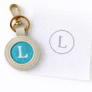 May include: A round keychain with a teal center embroidered with the letter "L" in white. The keychain has a light gray leather border and a gold-colored clasp. A white fabric square with a gray "L" design is next to the keychain.