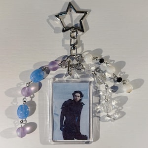 May include: A clear acrylic keychain featuring a photo of a person in dark clothing. The keychain is adorned with a silver star charm, silver chain links, and beaded strands in blue, purple, white, and black. The beads are various shapes and sizes.