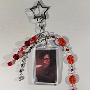 May include: A clear acrylic keychain featuring a portrait. The keychain is adorned with red and clear beads, and a silver star-shaped charm. The beads are strung on silver chains. The portrait is of a person with dark hair.