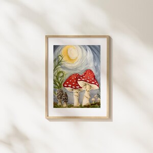 May include: A framed watercolor painting depicting two large red mushrooms with white spots, two smaller brown morel mushrooms, and green foliage under a yellow crescent moon against a blue sky. The artwork is presented in a light wood frame.