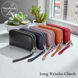 May include: A selection of long wristlet clutches in colours such as black, navy, mauve, orange, and red. Each clutch features a silver zip and a matching wrist strap. The image also includes a succulent and a small plant.