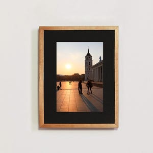 Puede incluir: Una fotografía enmarcada de una ciudad con un alto edificio de iglesia blanco en el fondo. La foto fue tomada al atardecer, y el cielo es de un naranja y rosa vibrantes. Hay personas caminando en primer plano, y el suelo está pavimentado con adoquines.