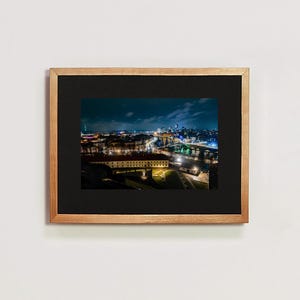 Puede incluir: Una vista aérea nocturna de un horizonte urbano con un río que lo atraviesa. La ciudad está iluminada con luces, y hay edificios de diferentes alturas. El río también está iluminado, y hay puentes que lo cruzan. La imagen está enmarcada en un marco de madera.