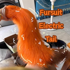 以下が含まれることがあります： オレンジと茶色のフェイクファーの尻尾。おそらくファースーツ用。箱の前で尻尾が持ち上げられています。「Fursuit Electric Tail」の文字がオレンジ色で書かれています。鮮やかなアクセサリーです。