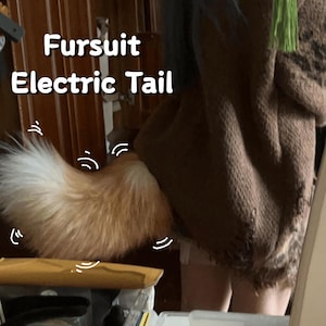 Puede incluir: Una cola de fursuit esponjosa, de color naranja claro y blanco. El texto "Fursuit Electric Tail" se muestra en blanco y en negrita. La cola está unida a una prenda marrón.