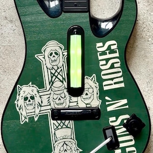 May include: Green guitar-shaped game controller with the text "GUNS N' ROSES" in white. The controller features a cross design with skull graphics. The controller has a black neck and a green light bar.