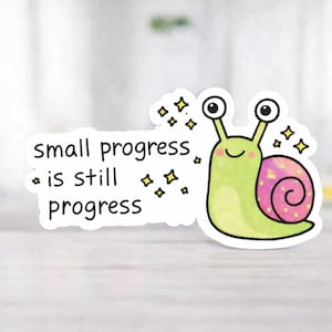 Könnte beinhalten: Aufkleber mit einer Cartoon-Schnecke mit grünem Körper und rosa-lila Gehäuse. Der Text "small progress is still progress" steht auf weißem Hintergrund. Gelbe Sterne umgeben die Schnecke.
