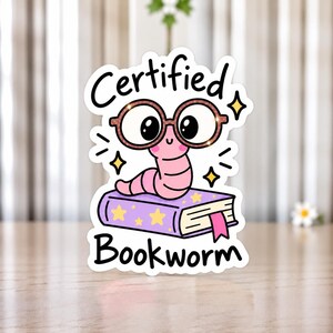 Peut inclure: Un autocollant découpé représentant un ver de terre rose en dessin animé portant de grandes lunettes marron, posé sur un livre violet décoré d'étoiles jaunes et d'un marque-page rose. Le texte "Certified Bookworm" est imprimé en noir.