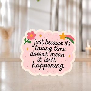 May include: A scalloped-edge sticker with a light pink background featuring small dots, a pink flower, a yellow star, and the text: just because it's taking time doesn't mean it isn't happening. The design includes a green stem and a pink swoosh.