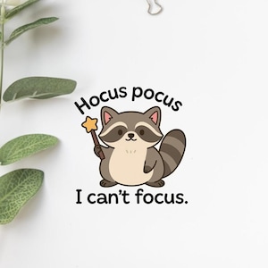 Könnte beinhalten: Ein Cartoon-Waschbär-Aufkleber mit dem Text "Hocus pocus" oben und "I can't focus" unten. Der Waschbär ist grau, braun und hellbraun und hält einen goldenen Sternenstab. Auf weißem Hintergrund.