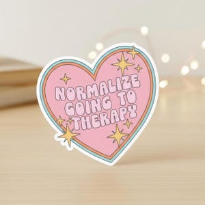 Könnte beinhalten: Ein herzförmiger Aufkleber mit dem Text "NORMALIZE GOING TO THERAPY" in einer Retro-Schriftart. Der Aufkleber hat eine rosa Mitte, einen hellblauen und orangefarbenen Rand und ist mit gelben Sternen verziert. Der Aufkleber befindet sich auf einer Holzoberfläche.