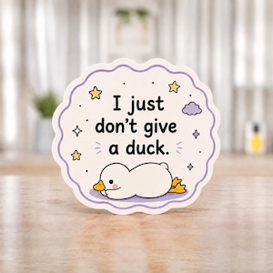 Könnte beinhalten: Ein weißer, kreisförmiger Aufkleber mit gewelltem Rand, der eine weiße Cartoon-Ente zeigt, die flach auf dem Bauch liegt. Der Text lautet, I just don't give a duck. Das Design enthält kleine gelbe Sterne, eine lila Wolke und Funkeln.