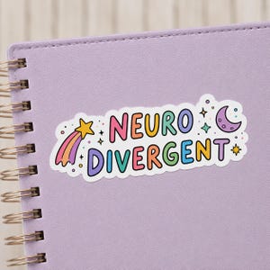 Könnte beinhalten: Ein gestanzter Aufkleber mit dem Text NEURO DIVERGENT in bunten, abgerundeten Buchstaben. Das Design enthält einen gelben Sternschnuppen-Akzent mit violettem Schweif links, einen violetten Halbmond rechts sowie kleine bunte Sterne und Punkte.