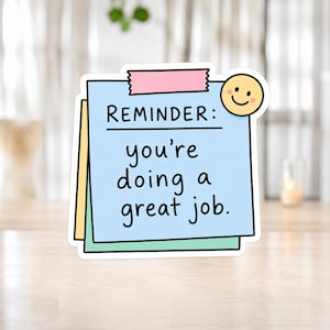Könnte beinhalten: Ein quadratischer Aufkleber mit einem hellblauen Haftnotizzettel mit dem Text REMINDER: you're doing a great job. Der Zettel ist mit einem rosa Klebestreifen und einem kleinen gelben Smiley-Symbol versehen und liegt auf einem gelben und grünen Zettel.