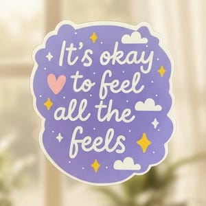Könnte beinhalten: Ein lila Aufkleber mit dem Schriftzug "It's okay to feel all the feels" in Weiß. Der Aufkleber hat einen wolkenförmigen Rand, ein rosa Herz, gelbe Sterne und weiße Wolken.
