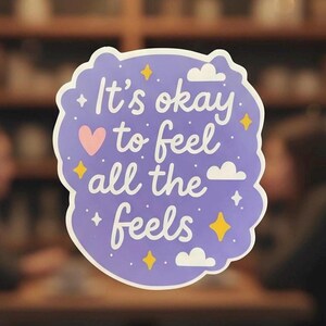 May include: A sticker with a light purple background and a white border. The sticker has the text "It's okay to feel all the feels" in white script. There is a pink heart, yellow stars, white clouds, and small white stars.