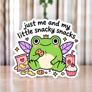 Könnte beinhalten: Ein gestanzter Aufkleber mit einem grünen Frosch mit lila Feenflügeln, einer kleinen Krone und rosa Wangen. Der Frosch isst einen Keks mit Schokostückchen, umgeben von einer Tüte Chips, einer Erdbeersaftpackung und einem Cupcake. Text: just me and my little snacky snacks.