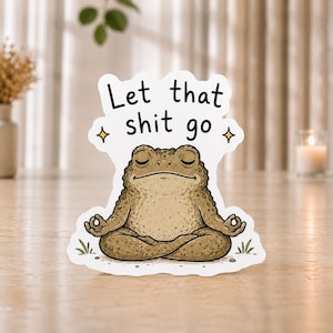 Könnte beinhalten: Ein gestanzter Aufkleber mit einer braunen, texturierten Kröte, die mit geschlossenen Augen in einer Meditationshaltung sitzt. Über der Kröte steht der schwarze Text Let that shit go, flankiert von zwei kleinen gelben Sternen.