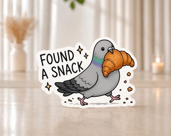 Könnte beinhalten: Ein gestanzter Aufkleber mit einer grauen Taube mit schillerndem Halsfleck, die ein goldbraunes Croissant im Schnabel hält. Der Text FOUND A SNACK ist in schwarzen Großbuchstaben neben dem Vogel gedruckt, ergänzt durch kleine Sternsymbole und Krümel.