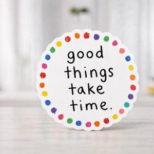 Könnte beinhalten: Ein weißer Aufkleber mit gewelltem Rand und dem Text "good things take time". Der Rand ist mit bunten Punkten in Rot, Orange, Gelb, Grün, Blau und Lila verziert. Der Aufkleber befindet sich auf einer hellen Oberfläche.