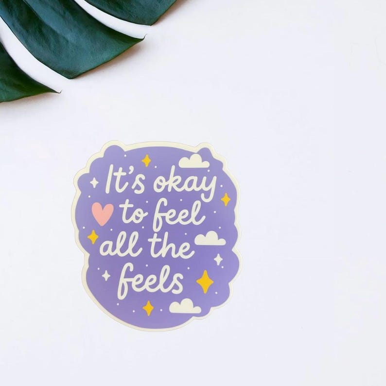 May include: A purple sticker with the phrase "It's okay to feel all the feels" in white script. The sticker has a cloud-like border, a pink heart, and yellow star accents. A green leaf is in the upper left corner.