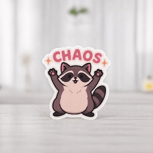 Puede incluir: Una pegatina con un mapache de dibujos animados con los brazos levantados. El mapache es marrón, beige y negro, con una barriga rosa. Encima del mapache está la palabra "CHAOS" en letras rosas con estrellas naranjas.