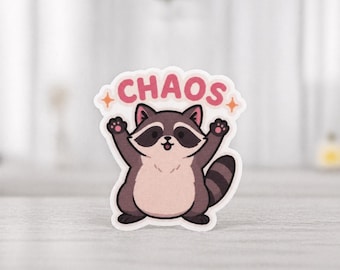 Könnte beinhalten: Ein Aufkleber mit einem Cartoon-Waschbären, der die Arme hochhält. Der Waschbär ist braun, hellbraun und schwarz, mit einem rosa Bauch. Über dem Waschbären steht das Wort "CHAOS" in rosa Buchstaben mit orangefarbenen Sternen.