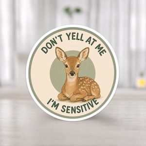 Könnte beinhalten: Ein runder Aufkleber mit einer Aquarellillustration eines Rehs und dem Text "DON'T YELL AT ME I'M SENSITIVE". Das Reh ist braun mit weißen Flecken, liegt vor einem grünen Kreis auf cremefarbenem Hintergrund.