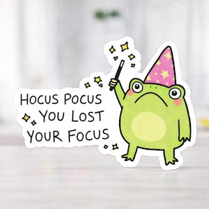 Könnte beinhalten: Ein Aufkleber mit einem Cartoon-Frosch, der einen Zaubererhut mit rosa Sternen trägt. Der Frosch hält einen Zauberstab und der Text lautet "Hocus Pocus, You Lost Your Focus". Der Frosch ist grün mit weißem Umriss, die Sterne sind gelb.