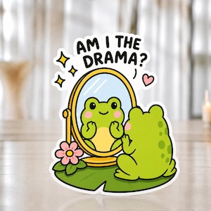 May include: A die-cut sticker featuring a green frog sitting on a lily pad, looking into a gold oval mirror. The text AM I THE DRAMA? is printed above the mirror, accompanied by yellow star icons and a small pink heart. The frog has pink cheeks.
