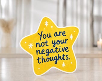Könnte beinhalten: Ein sternförmiges gelbes Schild mit weißem Rand und dem dunkelblauen Text "You are not your negative thoughts." Das Design enthält kleine weiße Stern- und Punktakzente, die über die Oberfläche des gelben Sterns verteilt sind.