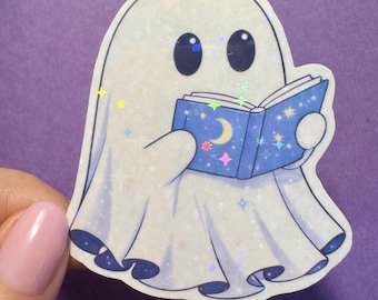 Könnte beinhalten: Ein weißer Geisteraufkleber, der ein blaues Buch mit einem Mond- und Sternenmuster liest. Der Geist hat große schwarze Augen und hält das Buch mit beiden Händen. Der Aufkleber hat einen holografischen, glitzernden Effekt auf einem lila Hintergrund.