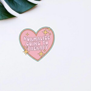 Puede incluir: Una pegatina en forma de coraz&oacute;n con el texto "Normalizar ir a terapia" en rosa, delineado en naranja y azul. La pegatina tiene acentos de estrellas y est&aacute; sobre un fondo blanco.