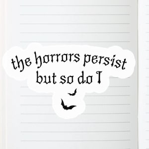 K&ouml;nnte beinhalten: Ein wei&szlig;er Aufkleber mit dem Text "the horrors persist but so do I" in gotischer Schrift. Zwei schwarze Fledermaus-Silhouetten befinden sich unter dem Text. Der Aufkleber befindet sich auf einer linierten Notizbuchseite.