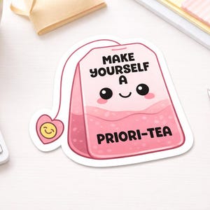 Könnte beinhalten: Ein rosa Teebeutel-Aufkleber mit dem Text "MAKE YOURSELF A PRIORI-TEA" in Schwarz. Der Teebeutel hat ein lächelndes Gesicht und ein herzförmiges Etikett mit einem lächelnden Gesicht. Der Aufkleber befindet sich auf einer weißen Oberfläche mit Tastatur und Stift.