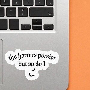 K&ouml;nnte beinhalten: Ein wei&szlig;er Aufkleber mit dem Text "the horrors persist but so do I" in gotischer Schrift, zusammen mit zwei Fledermaus-Silhouetten. Der Aufkleber befindet sich auf einem Laptop, mit einem orangefarbenen Hintergrund.