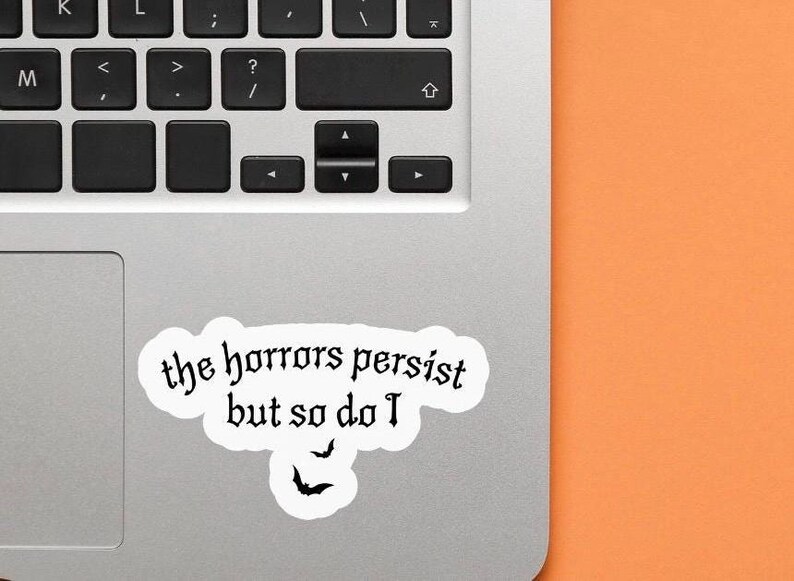 K&ouml;nnte beinhalten: Ein wei&szlig;er Aufkleber mit dem Text "the horrors persist but so do I" in gotischer Schrift, zusammen mit zwei Fledermaus-Silhouetten. Der Aufkleber befindet sich auf einem Laptop, mit einem orangefarbenen Hintergrund.