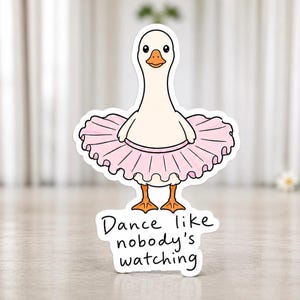 Könnte beinhalten: Ein Aufkleber mit einer Cartoon-Gans, die einen rosa Tutu trägt. Die Gans hat einen weißen Körper, einen orangefarbenen Schnabel und Füße sowie schwarze Augen. Der Aufkleber enthält den Text "Dance like nobody's watching".