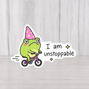 Op de afbeelding: Een sticker met een groene kikker op een roze fiets, met een feesthoed met roze en gele sterren. De tekst "I am unstoppable" staat naast de kikker, met sterren.