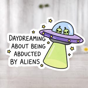Könnte beinhalten: Ein Aufkleber mit einer Cartoon-UFO mit zwei grünen Außerirdischen. Das UFO ist lila und silber, mit einem grünen Lichtstrahl. Der Text lautet "DAYDREAMING ABOUT BEING ABDUCTED BY ALIENS" mit gelben Sternakzenten.