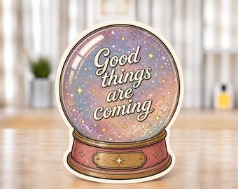 Puede incluir: Una pegatina troquelada con forma de bola de cristal con un interior brillante, iridiscente, morado y rosa. El texto 'Good things are coming' está impreso en una fuente cursiva oscura. La base es de color rosa apagado con una placa dorada.