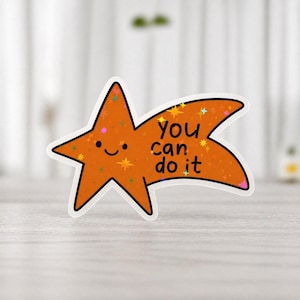 Op de afbeelding: Oranje ster-vormige sticker met de tekst "You can do it". De sticker heeft een witte rand, een lachend gezicht en kleurrijke steraccenten. De sticker ligt op een lichtgekleurd oppervlak.