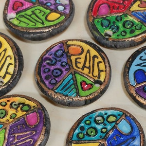 May include: A collection of colorful, hand-painted peace sign pendants. Each pendant is divided into three sections with the word "PEACE" in the center. The pendants have a dark border and a hole for stringing.