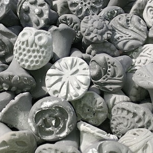 May include: A collection of gray and white ceramic stamps with various designs, including flowers, geometric patterns, and an owl. The stamps are arranged in a pile, showing the different designs and textures.