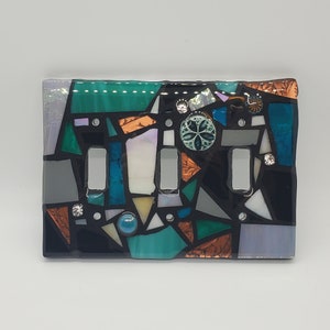 Pode incluir: Uma tampa de interruptor de luz de mosaico com fundo preto e uma variedade de cores, incluindo verde, azul, branco e cobre. A tampa do interruptor tem dois interruptores e um elemento decorativo no centro.